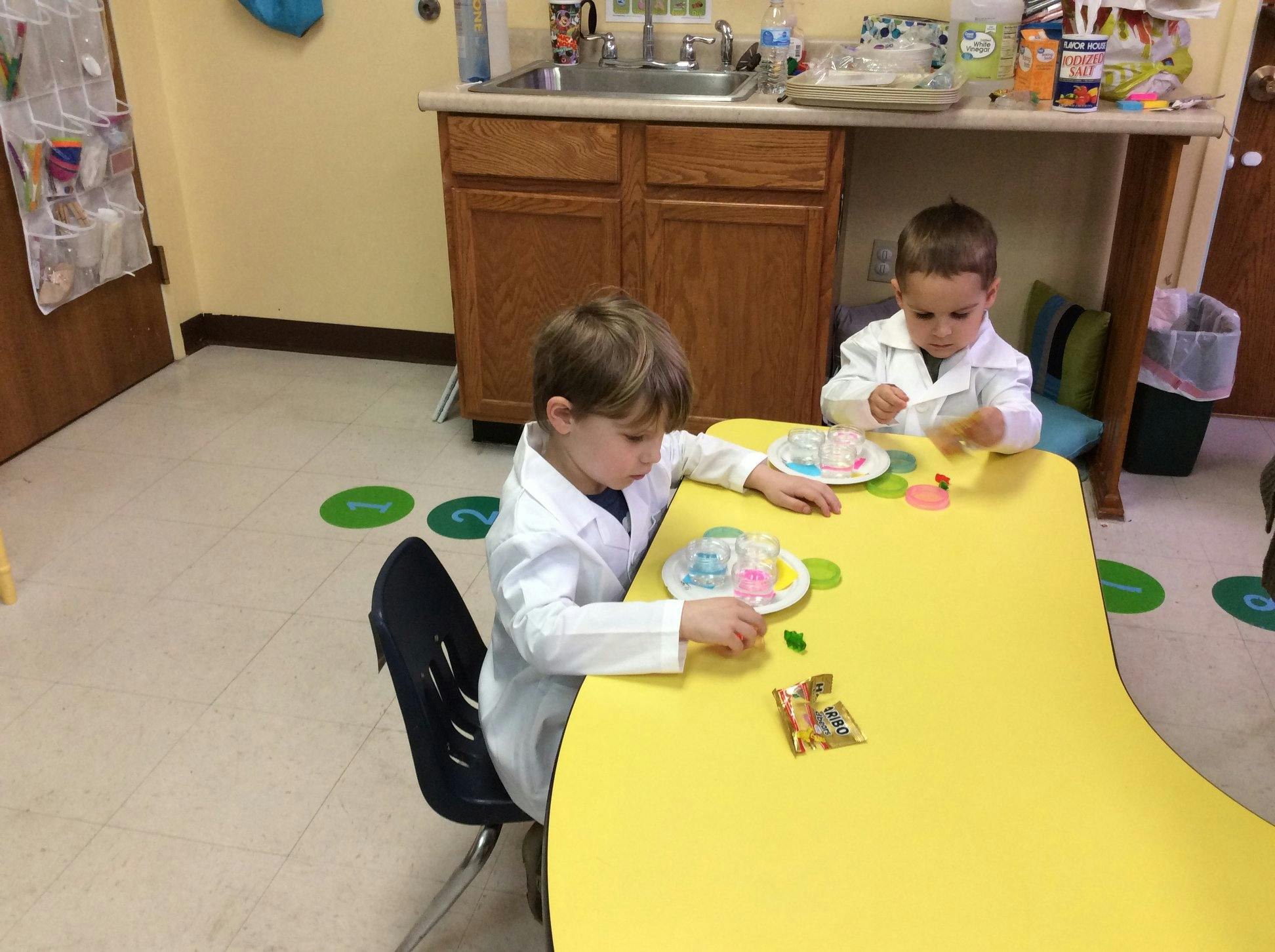Two children in lab coats playing with toys at a yellow table