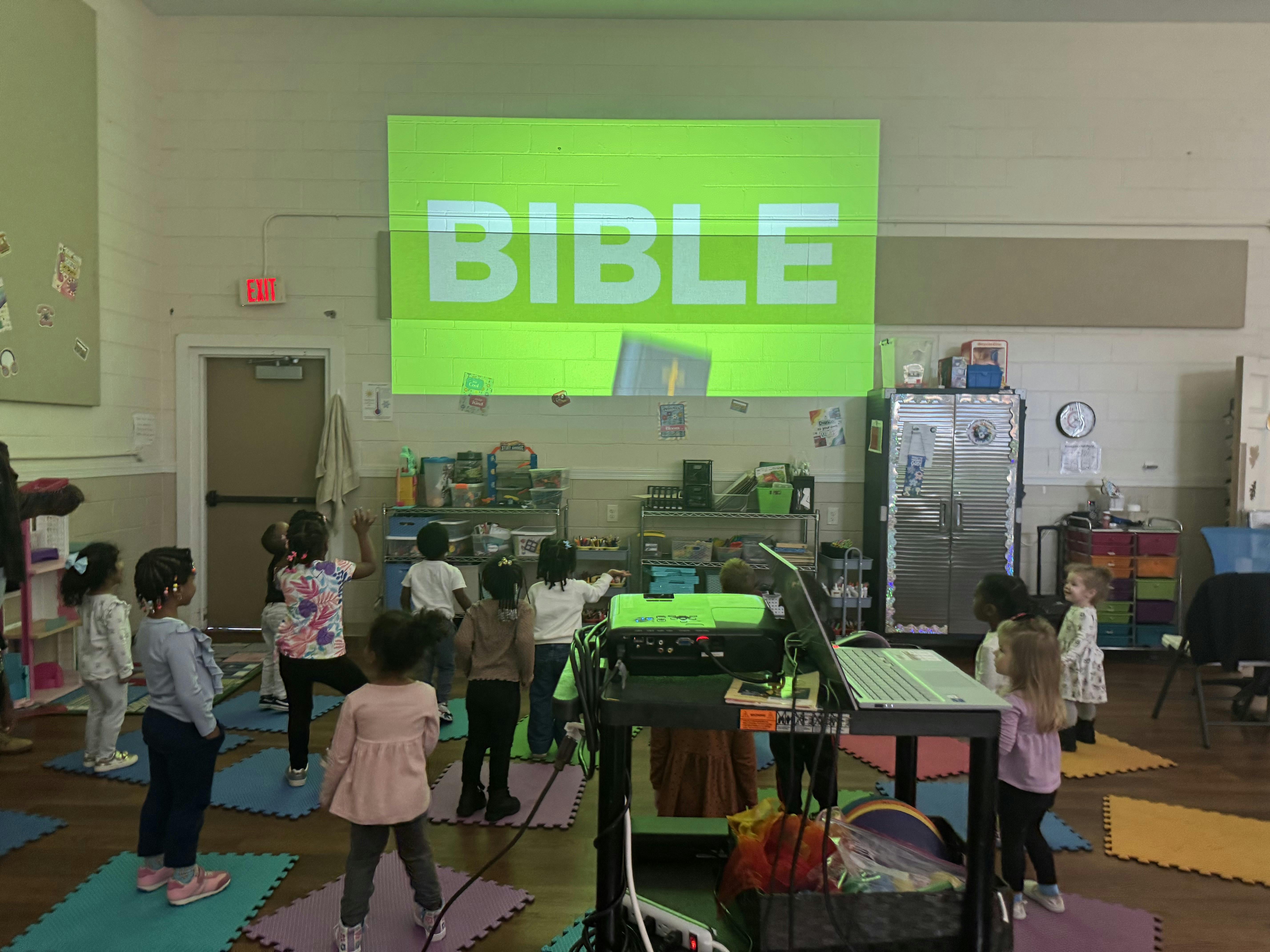 Group of children standing in a classroom facing a projected sign reading Bible