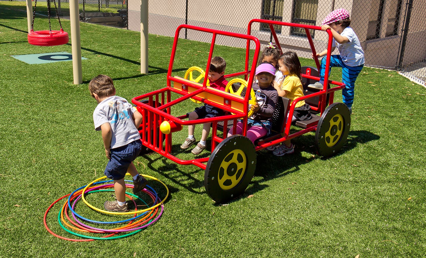 Children playing on a red toy car and with hula hoops on artificial grass