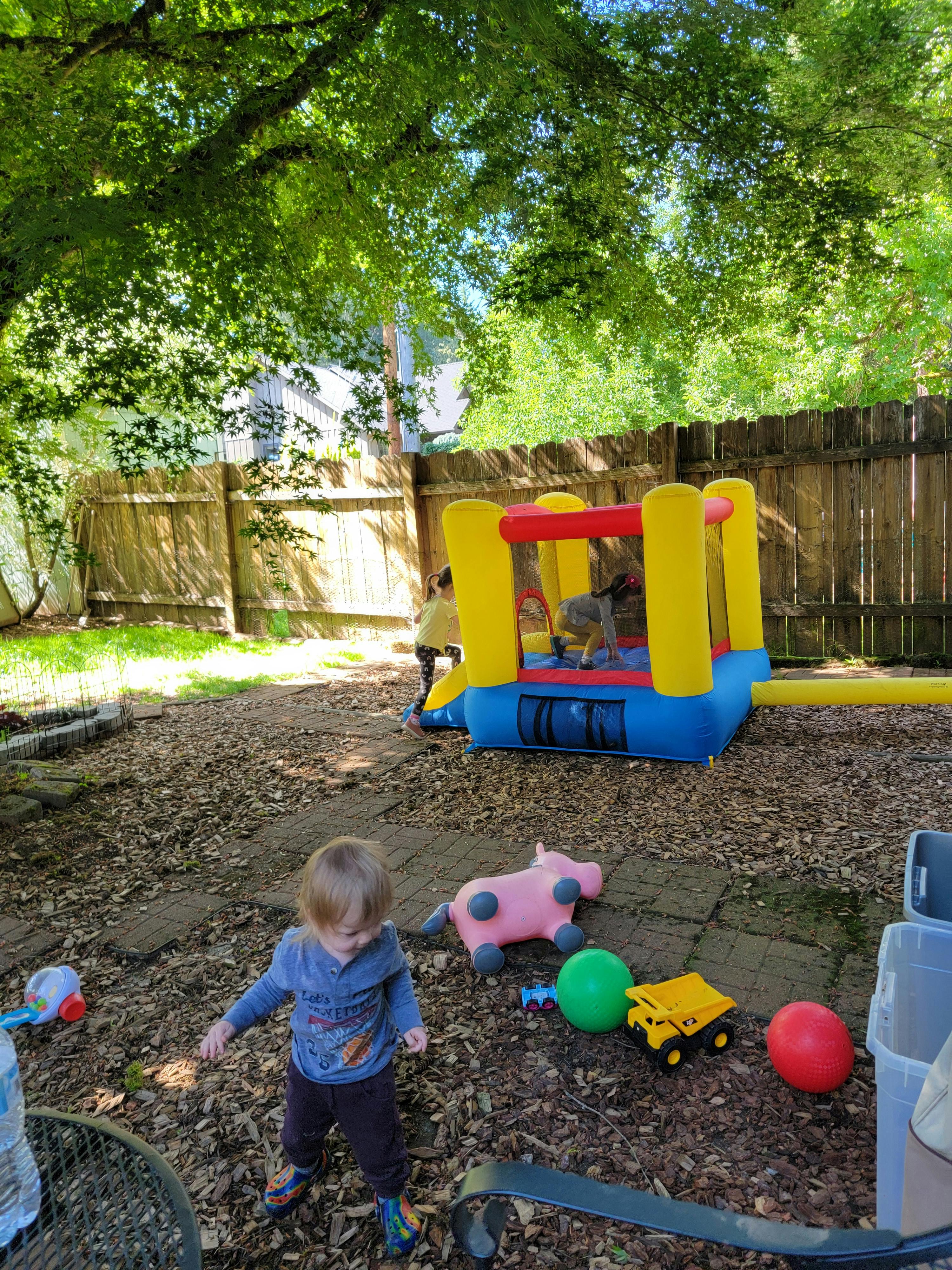 Child standing on wood chips with toys and bounce house in background