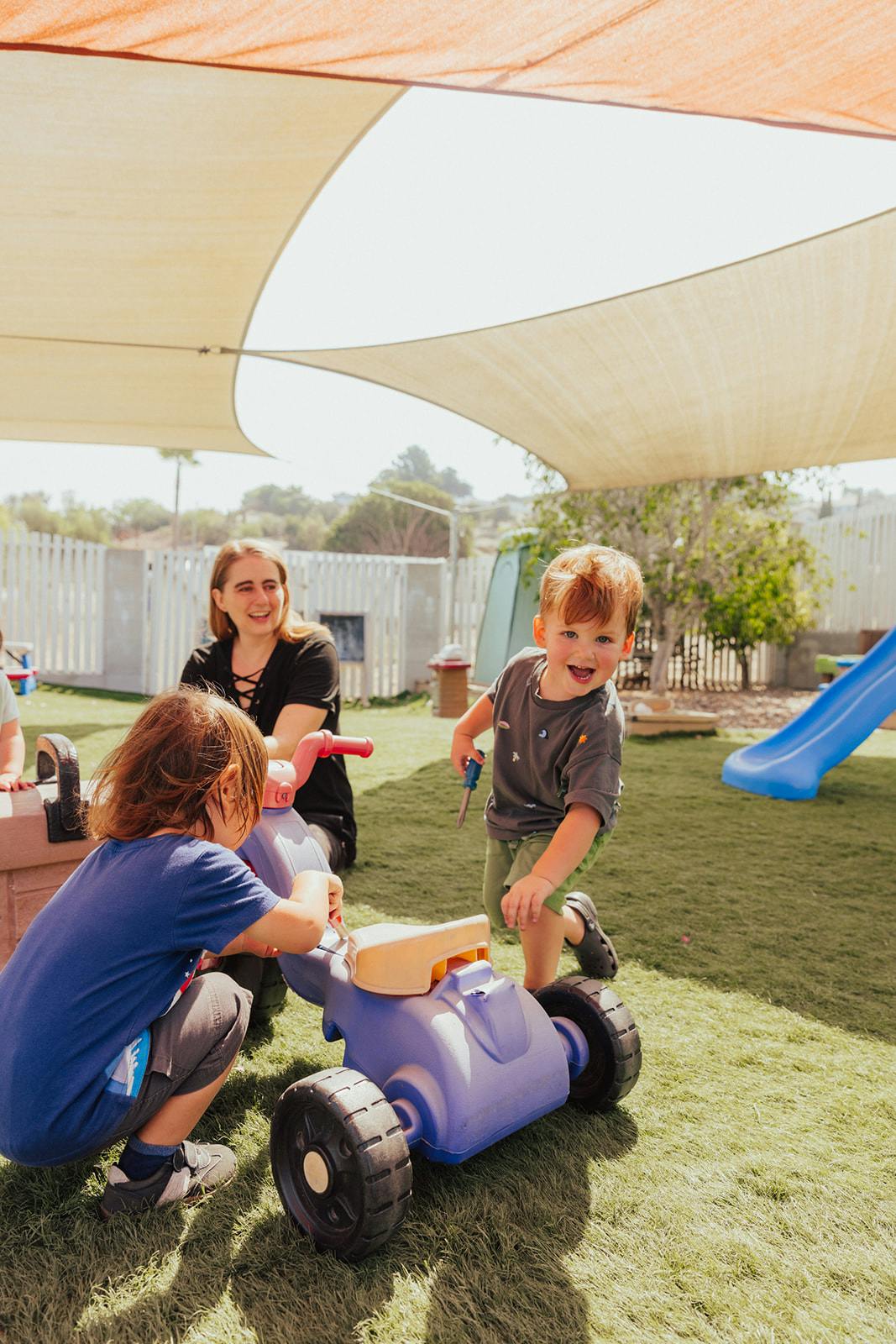 Two children playing with a toy car on grass, person in background