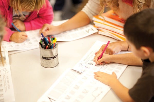 Two children and an adult writing on papers at a table with a can of markers