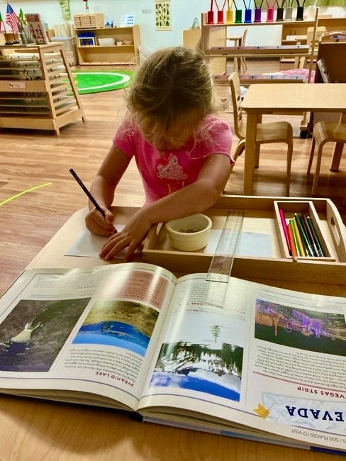 Child writing on paper at a desk next to an open book about Nevada