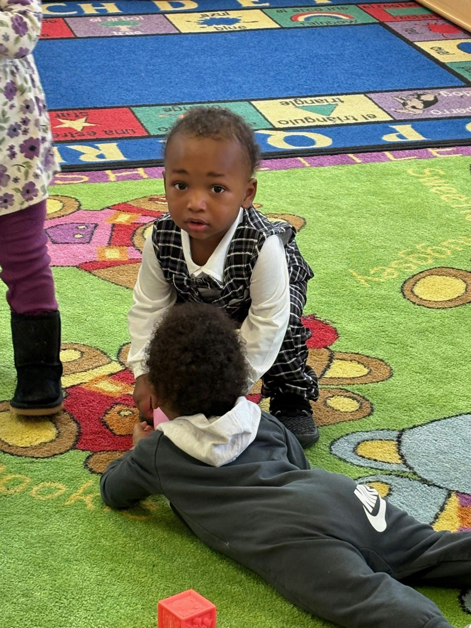 Child standing over another child on a colorful rug
