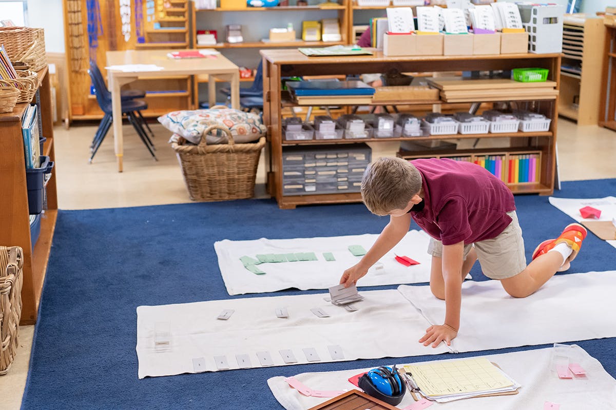 Child kneeling on floor, working with materials on a white mat in a classroom