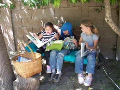 Three children sitting on a bench reading books outdoors