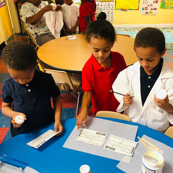 Three children at a table, one in a lab coat holding a brush