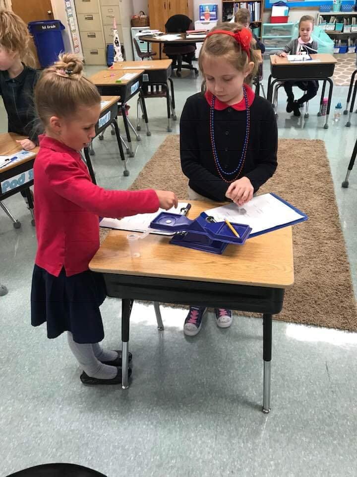 Two children at desks in a classroom, one standing and one seated