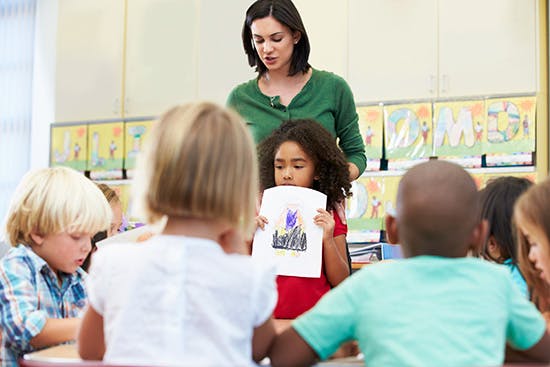 Adult and children in a classroom, child holding up a drawing