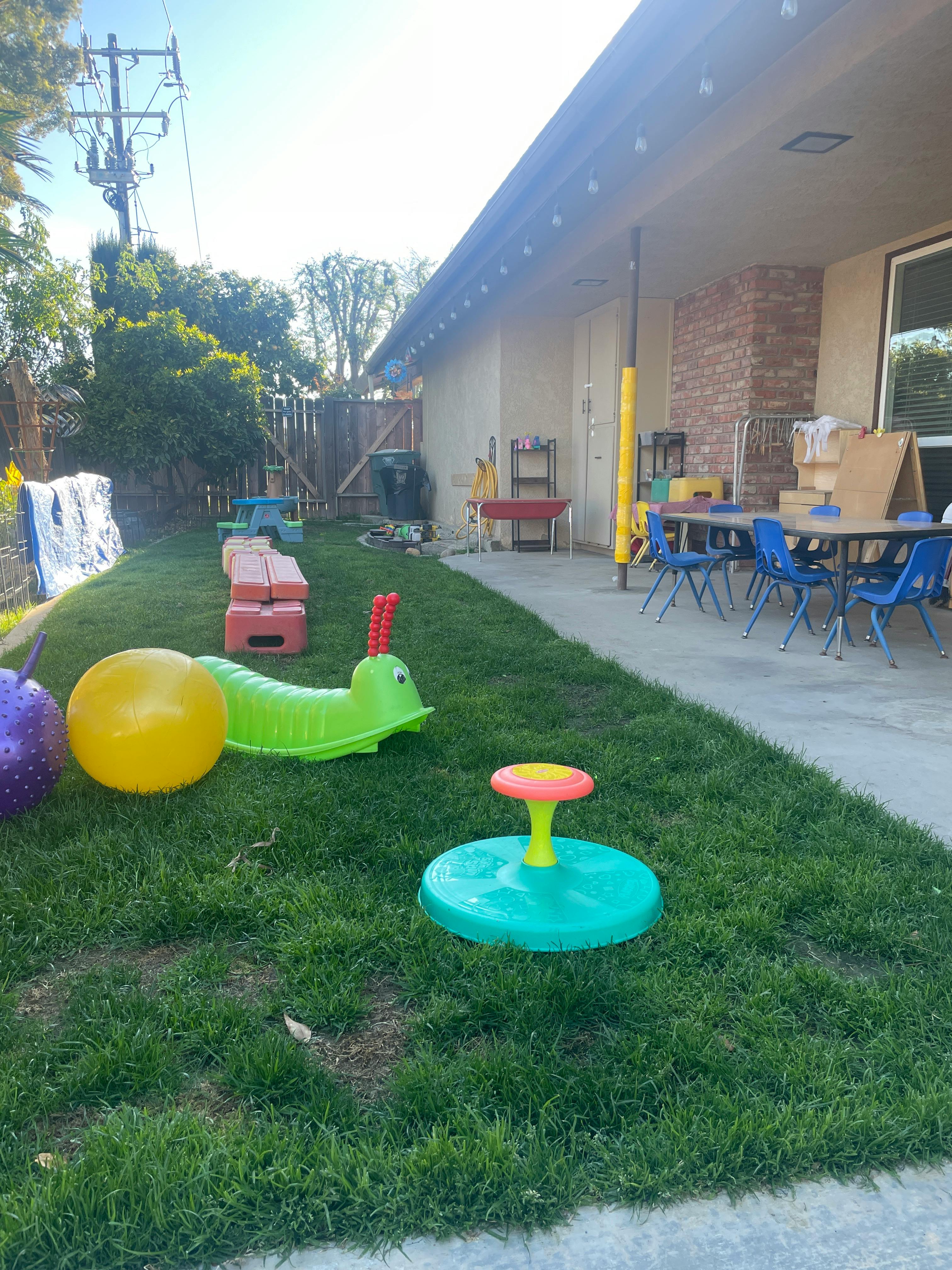 Outdoor play area with various toys on grass and tables with chairs on concrete