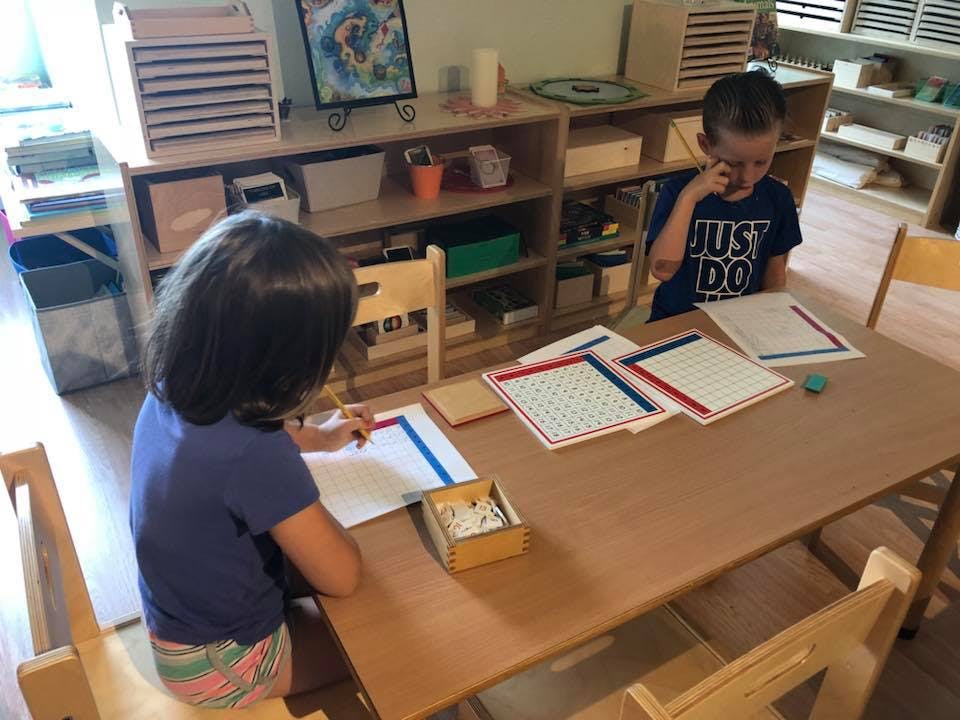 Two children at a table with papers and a box, one drawing