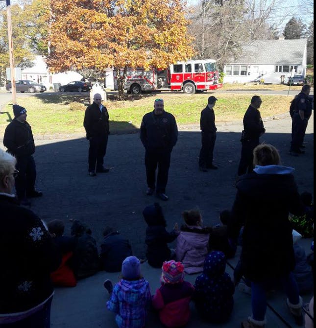 Adults standing and children sitting on the ground with a fire truck in the background