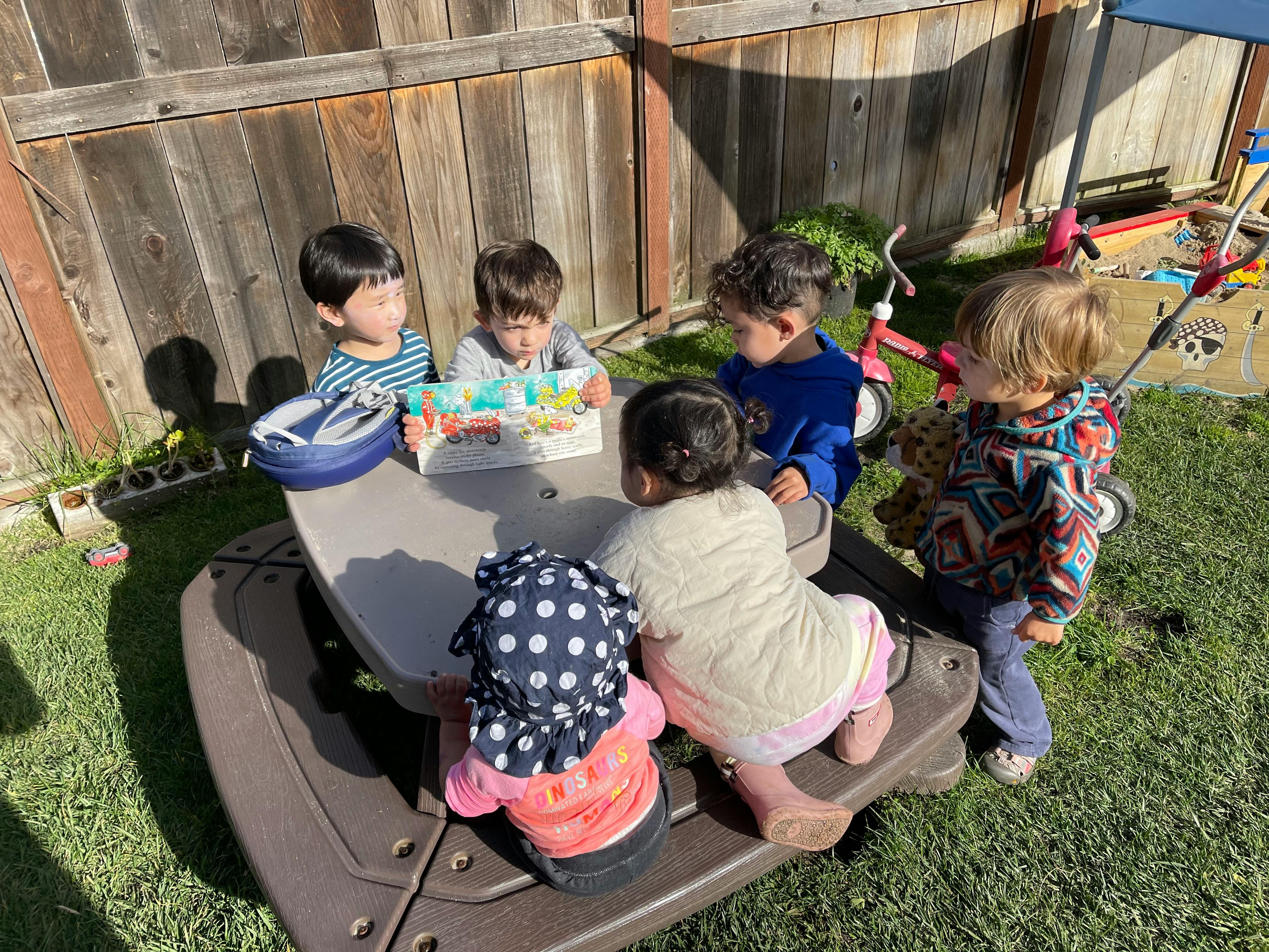 Five children gathered around a small picnic table outdoors