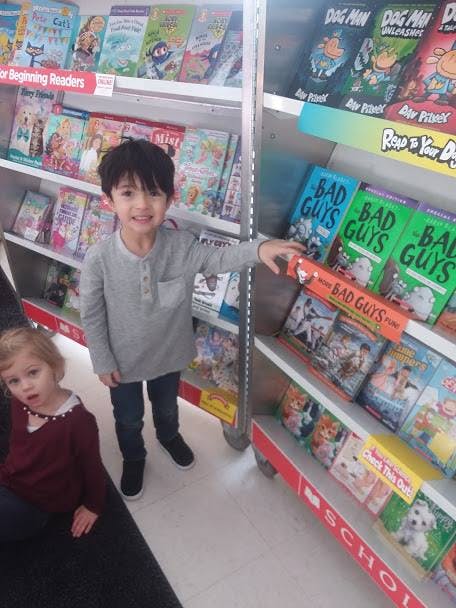 Child standing in front of a bookshelf, pointing at a book, with another child seated