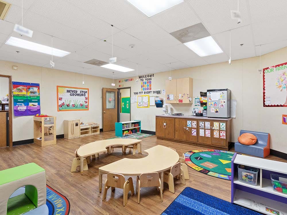 Classroom with small tables and chairs, and a counter with a refrigerator