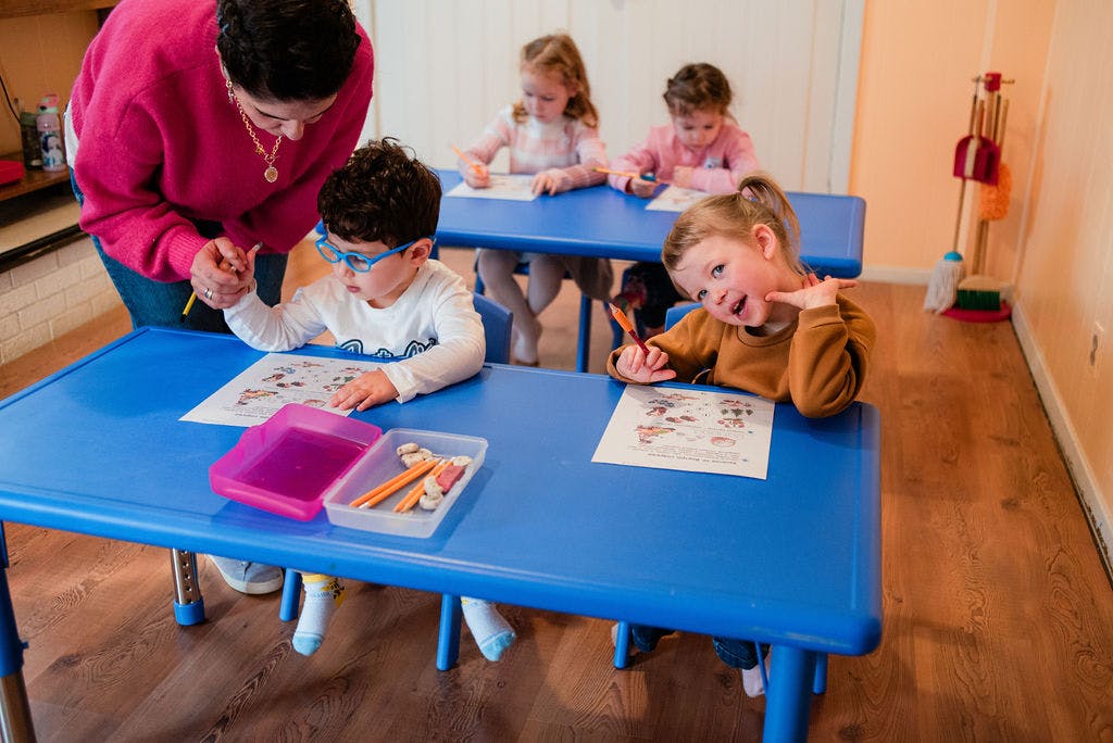 Adult helping a child color at a blue table with other children