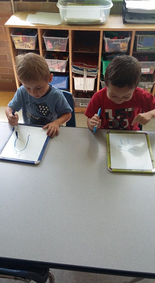 Two children drawing on whiteboards at a table in a classroom