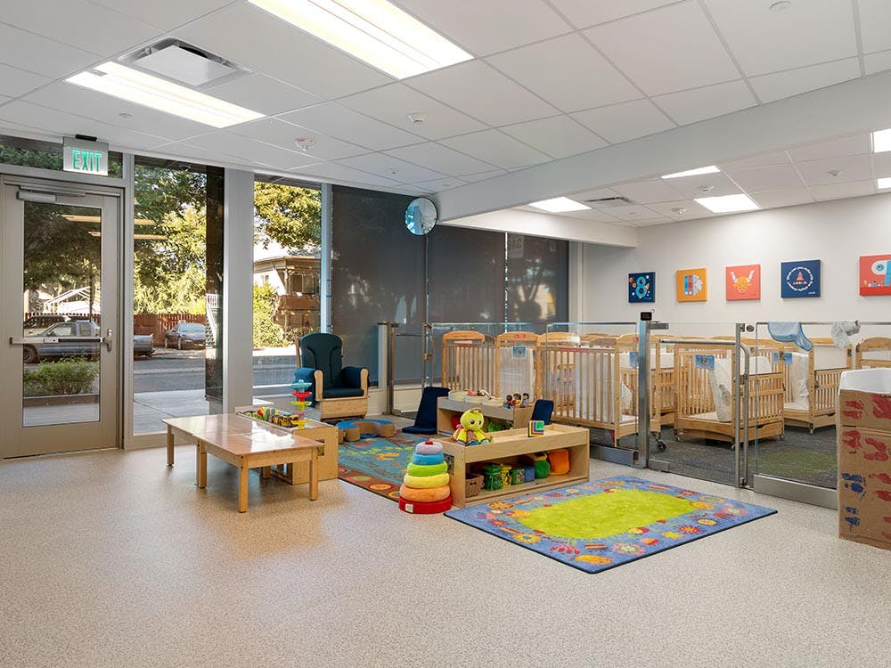 A childcare room with small cribs, play rugs, and an "EXIT" sign above a glass door