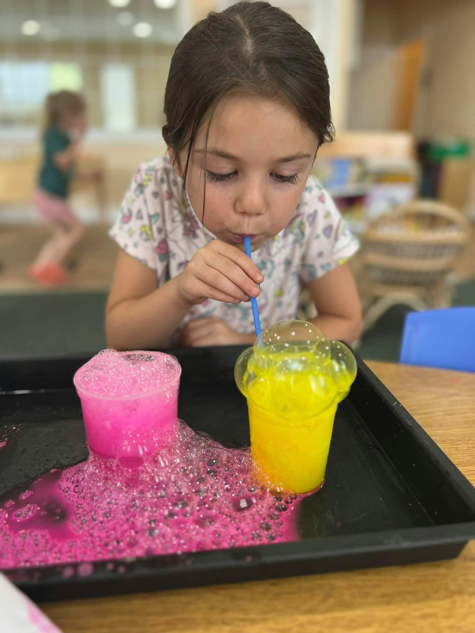 Child blowing bubbles into a yellow cup with a straw, pink bubbles overflowing from another cup