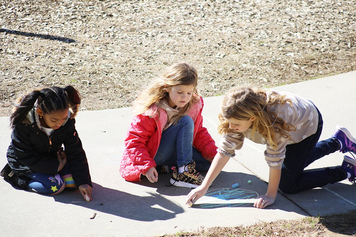 Three children kneeling on a sidewalk drawing with chalk