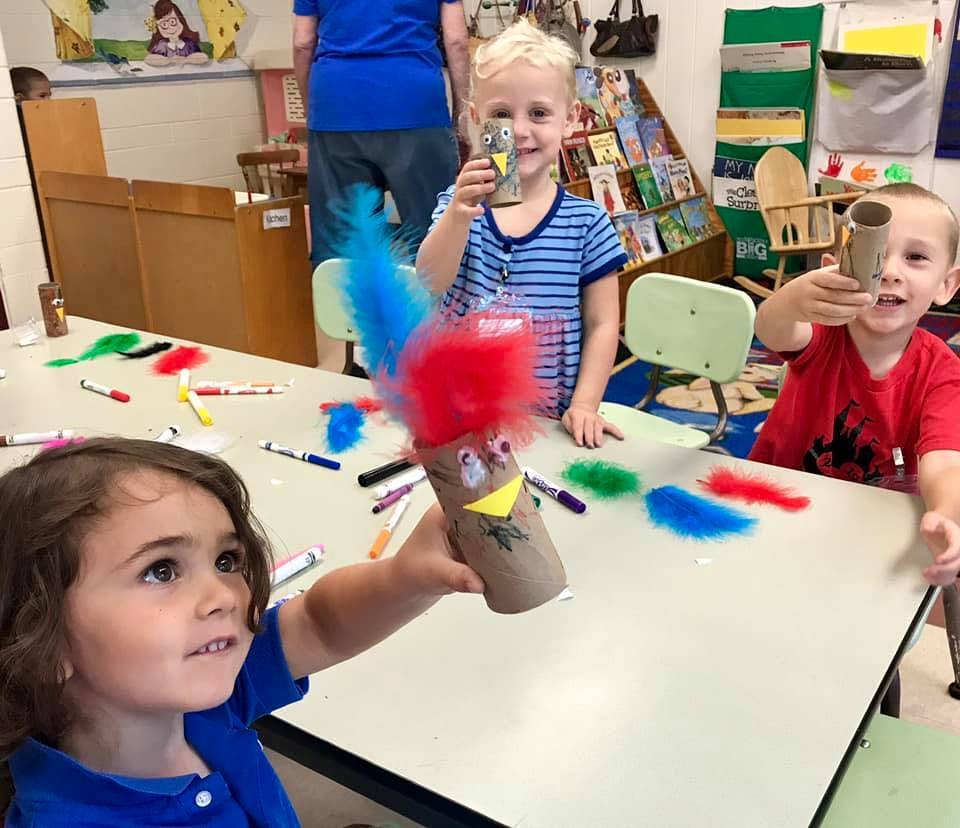 Children holding up crafts made from paper towel rolls and feathers in a classroom