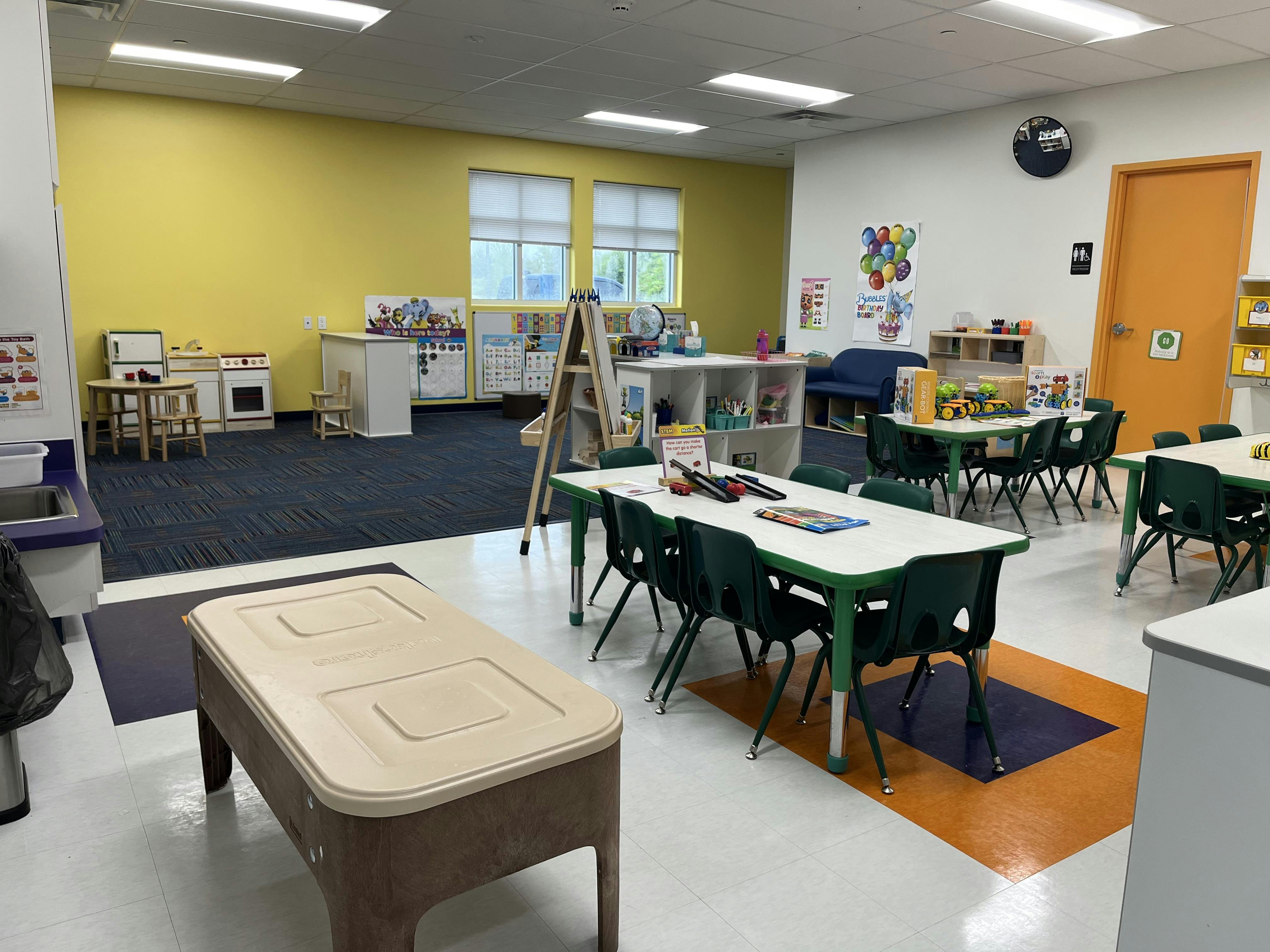 Classroom with tables and chairs, play kitchen, and yellow wall