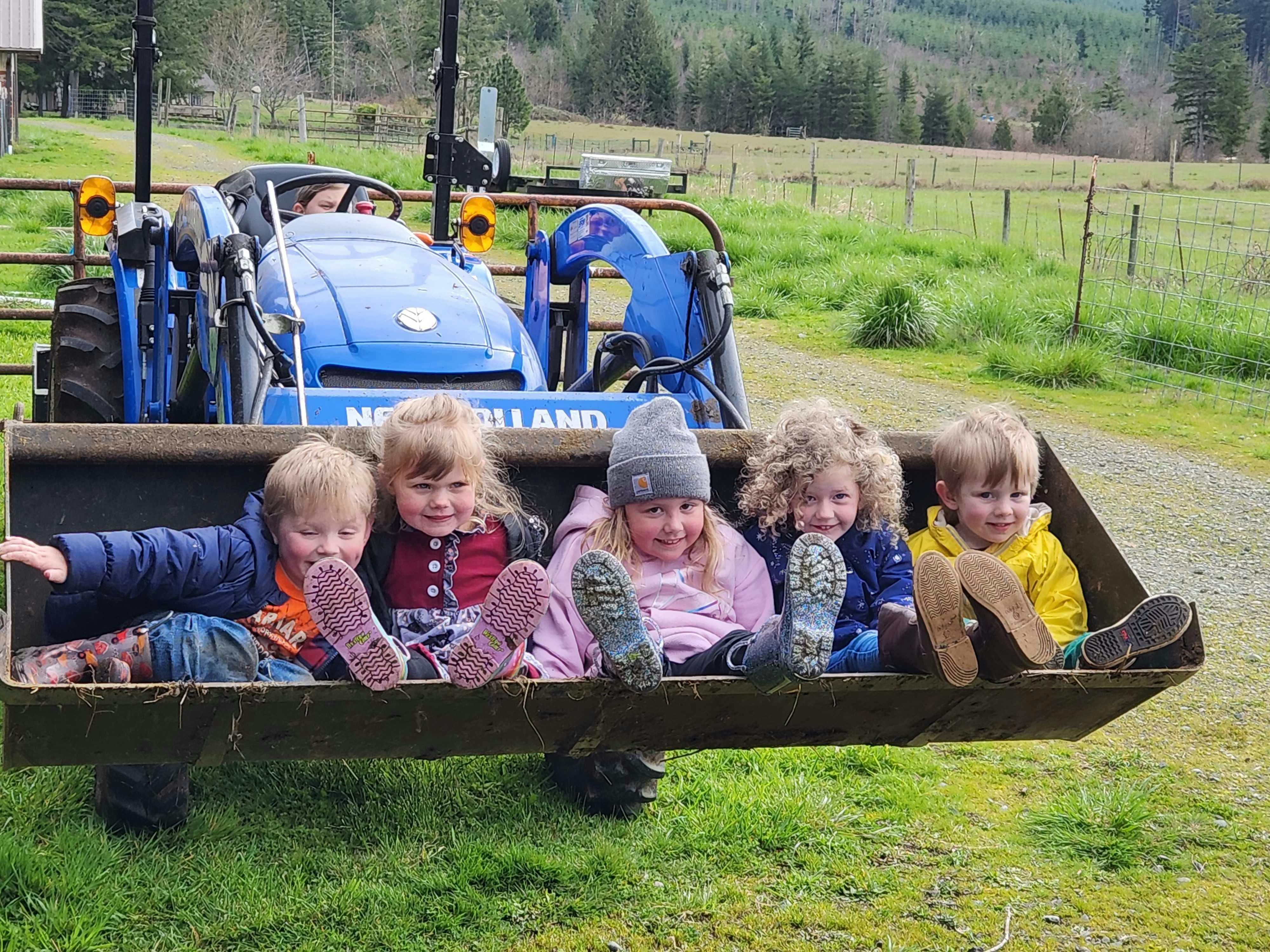 Five children sitting in the front loader bucket of a blue tractor