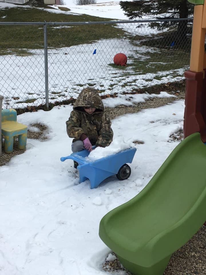 Child in a hooded jacket filling a blue toy wheelbarrow with snow
