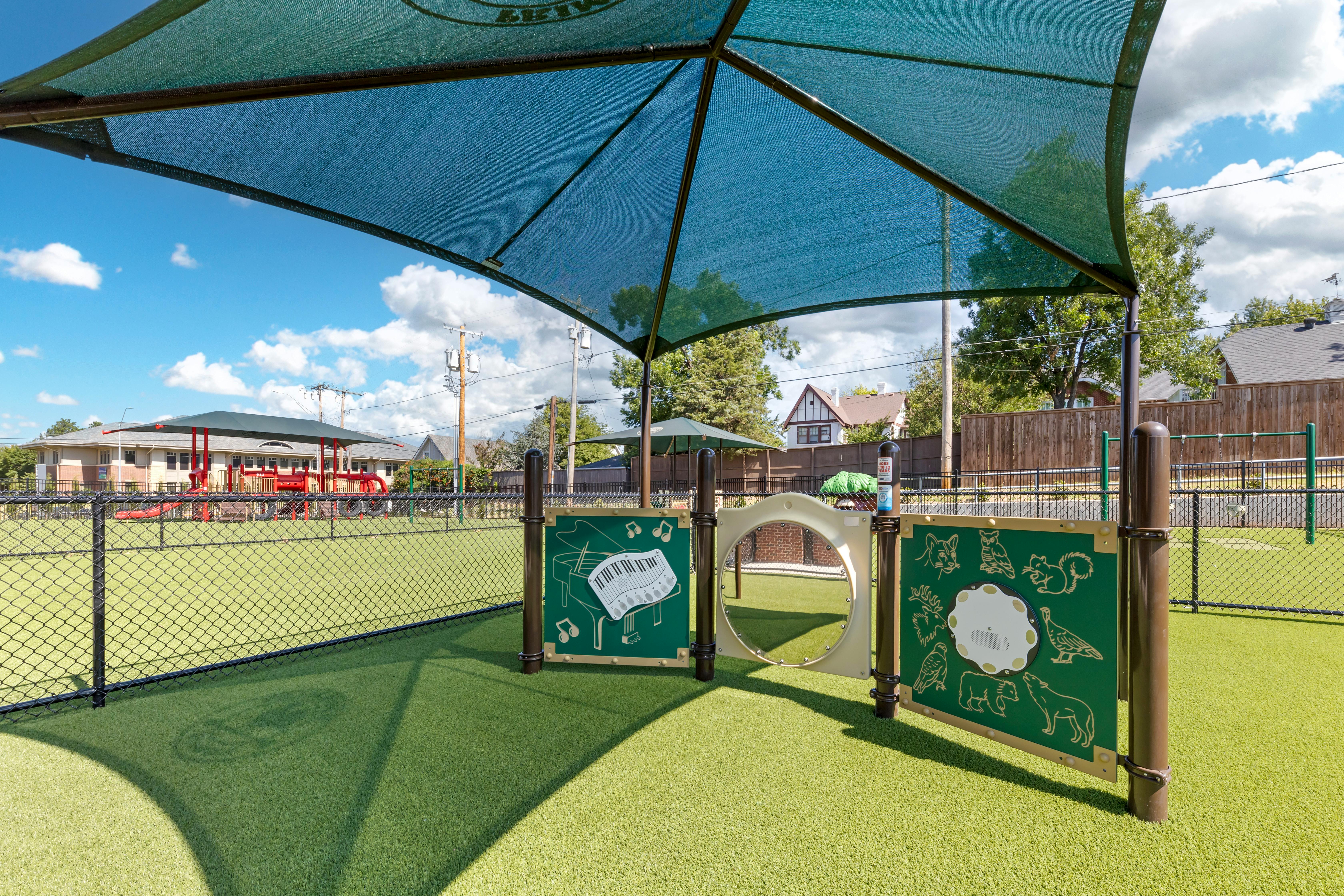 A green outdoor play area with artificial turf, a shade structure, and activity panels