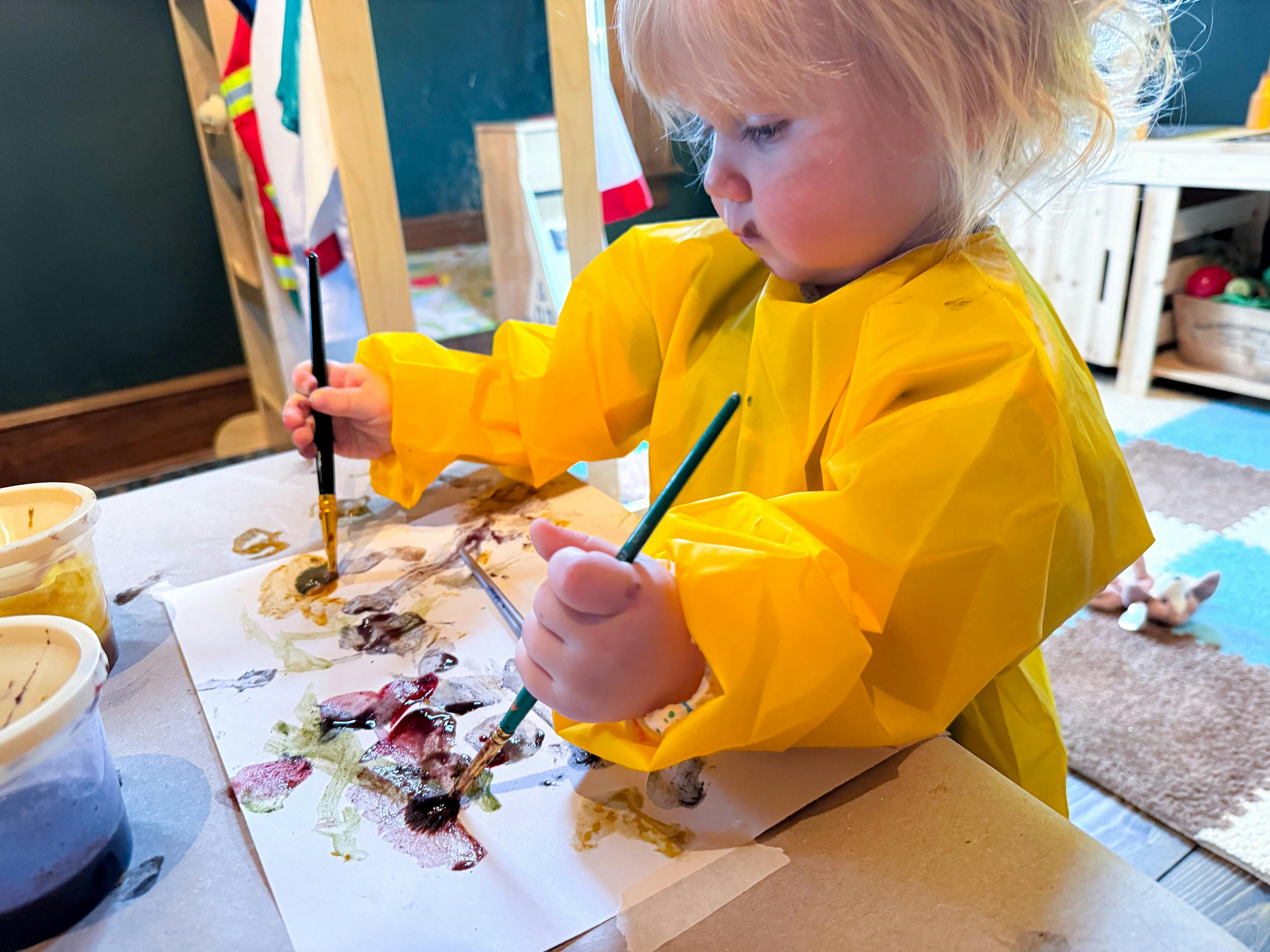 Child in a yellow smock painting with brushes on paper