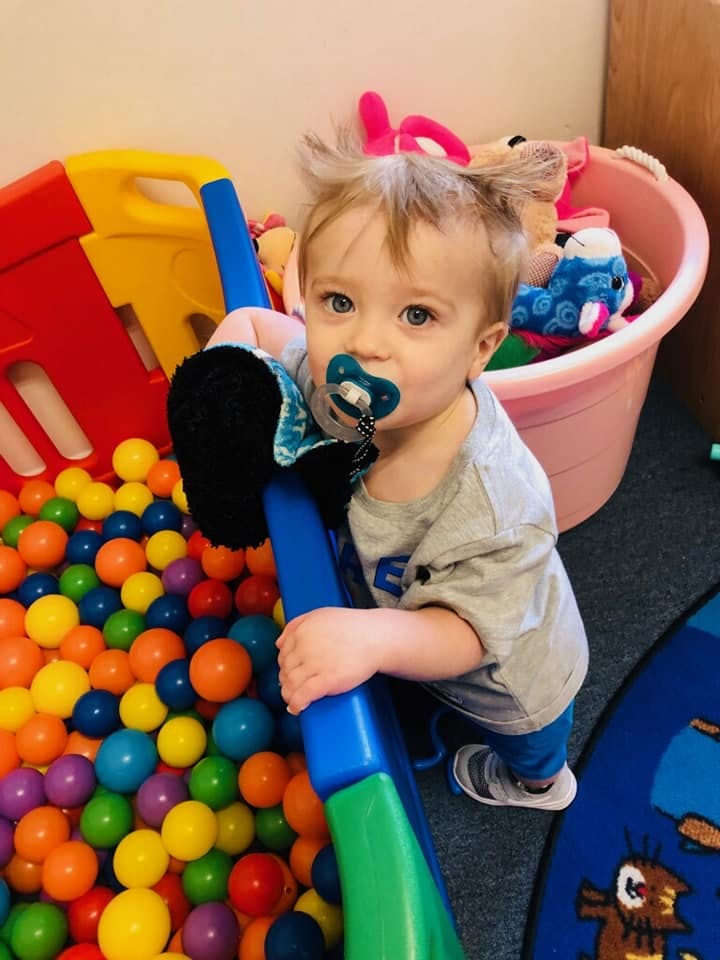 Child with a pacifier holding onto a blue and green toy next to a ball pit