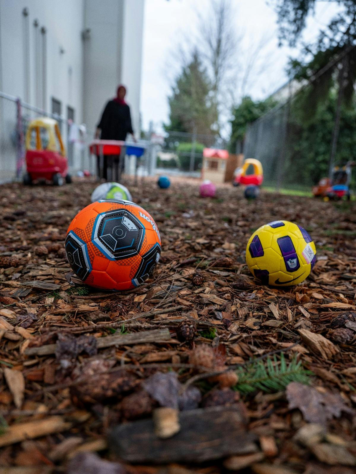 Two soccer balls on wood chips with a person and toys in the background
