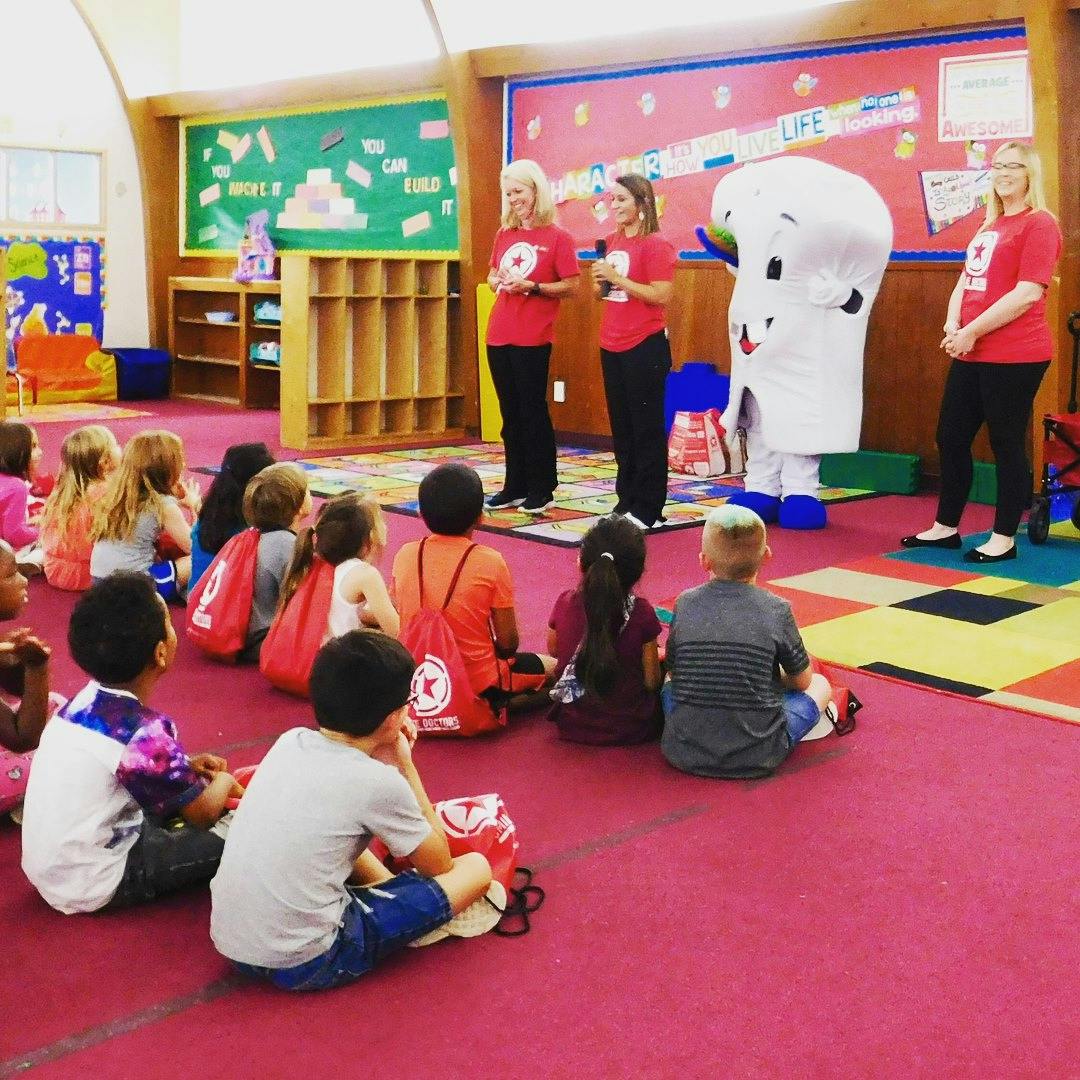 Children sitting on the floor watching adults and a mascot in a classroom