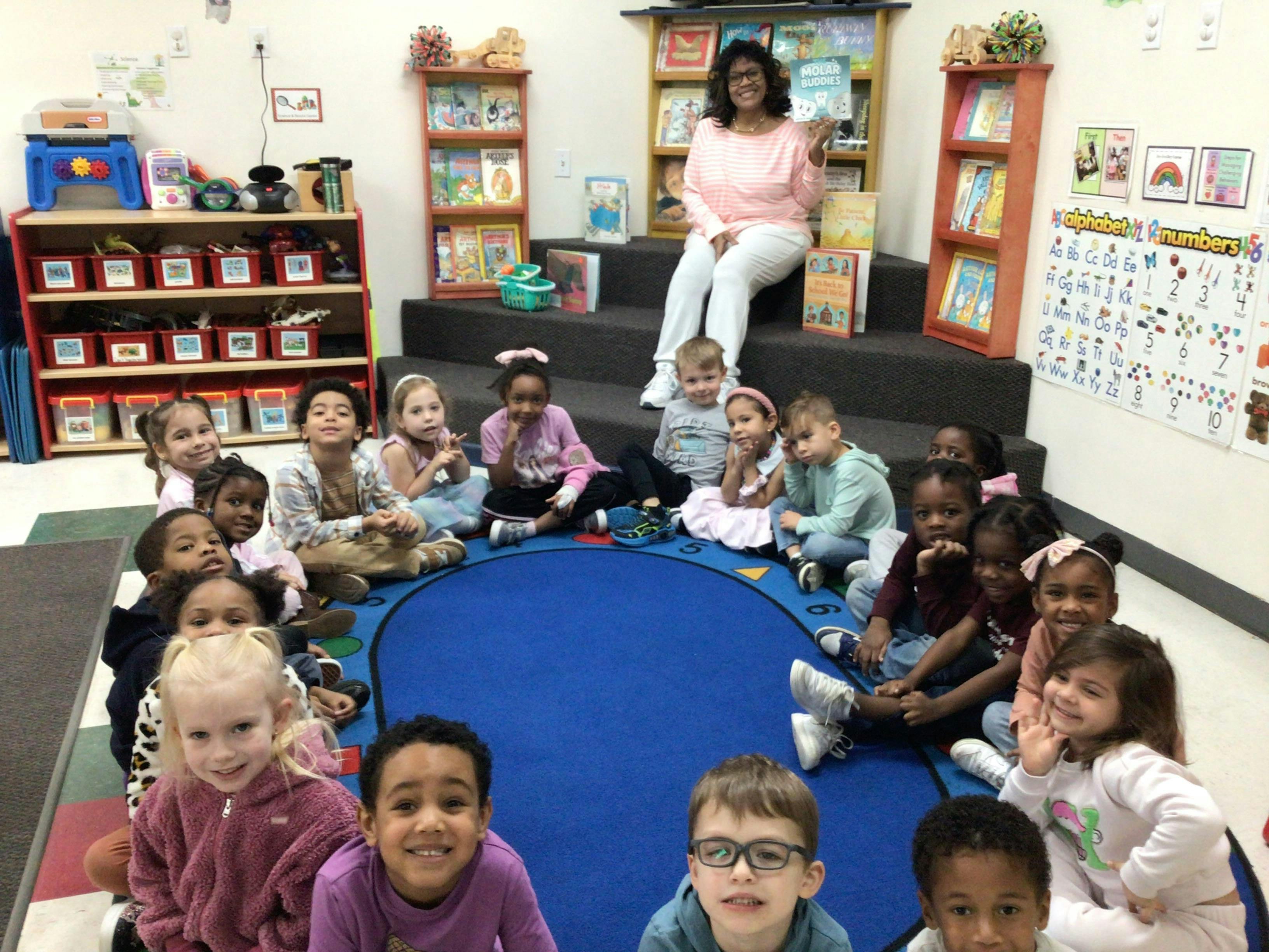 Group of children sitting on a classroom rug with an adult holding a book