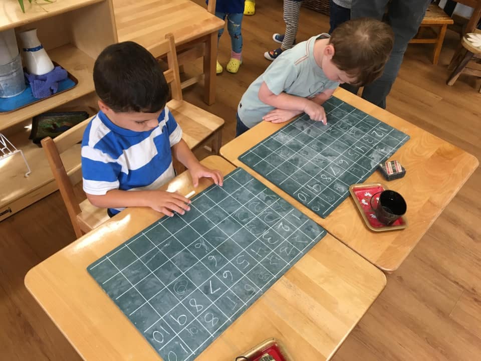 Two children writing on chalkboards at desks in a classroom
