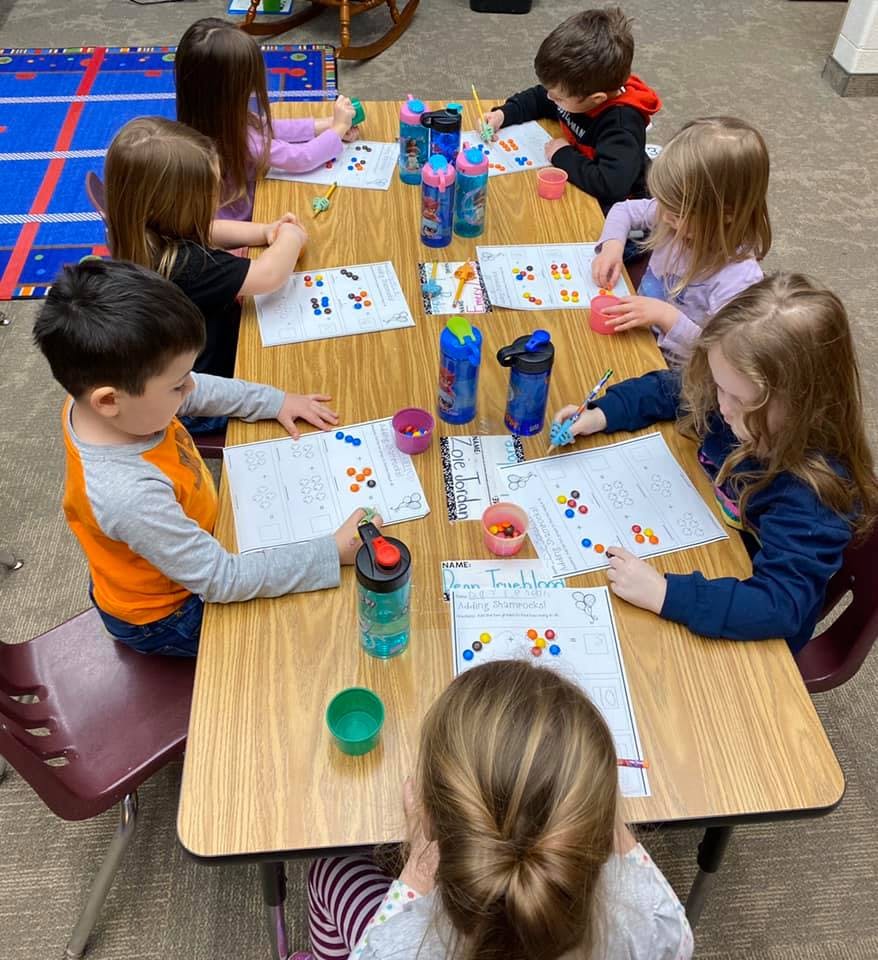 Children sitting at a table doing an activity with small colorful objects on papers