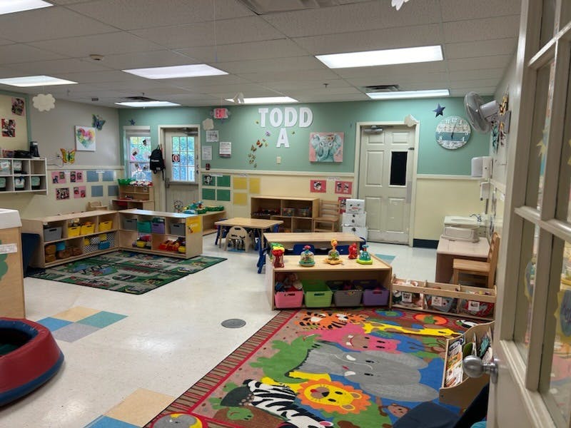 An empty classroom with colorful rugs, shelves of toys, and small tables and chairs