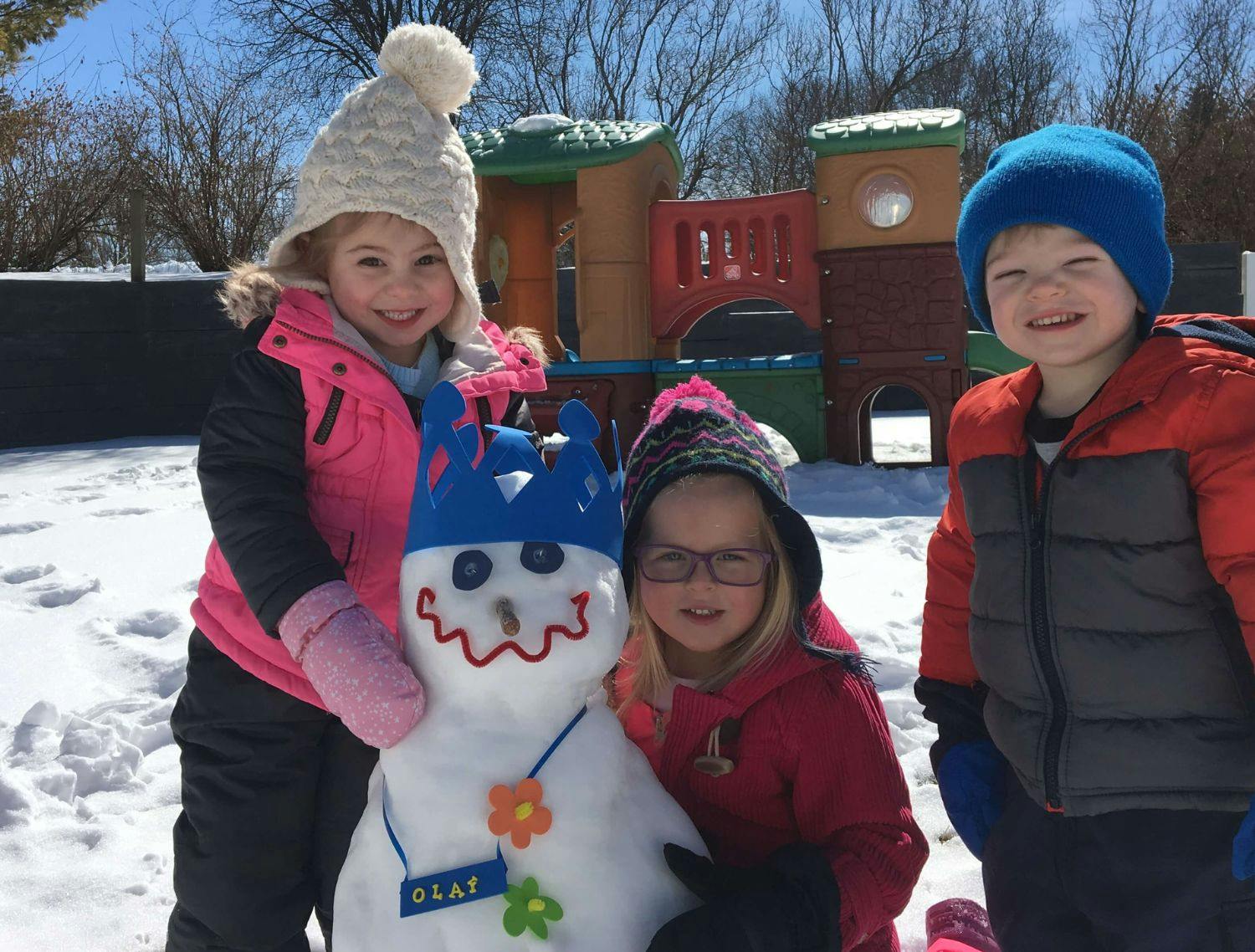 Three children standing in snow next to a snowman