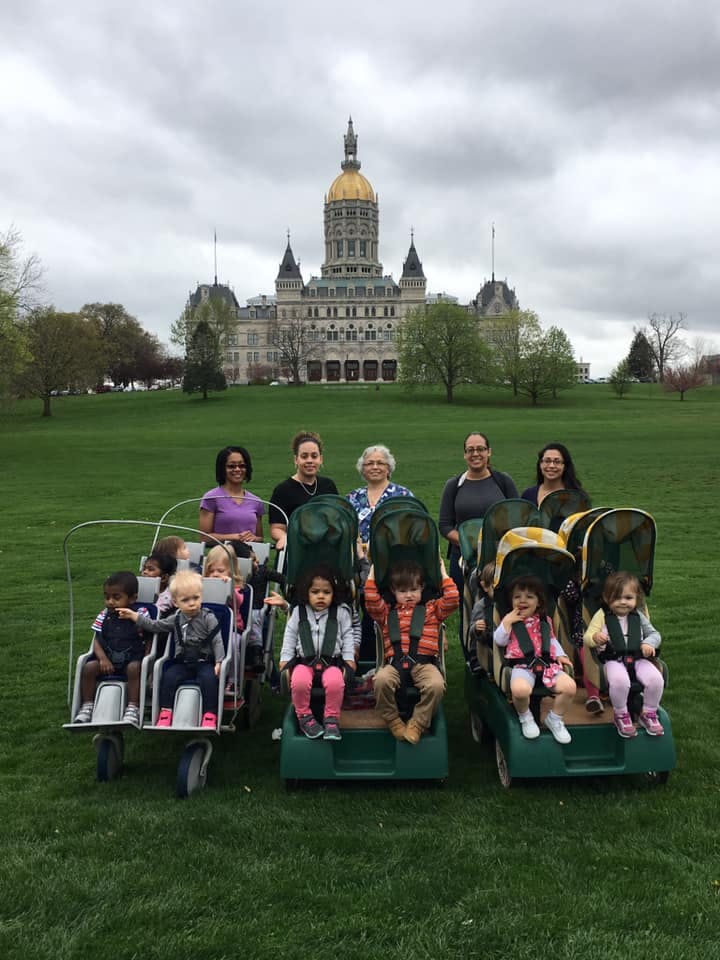 Adults and children in multi-child strollers on a lawn with a large building in the background