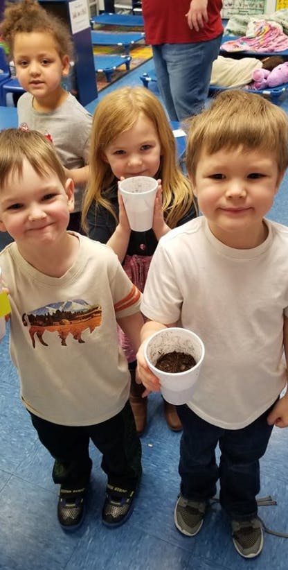 Three children holding cups with soil, one child in background