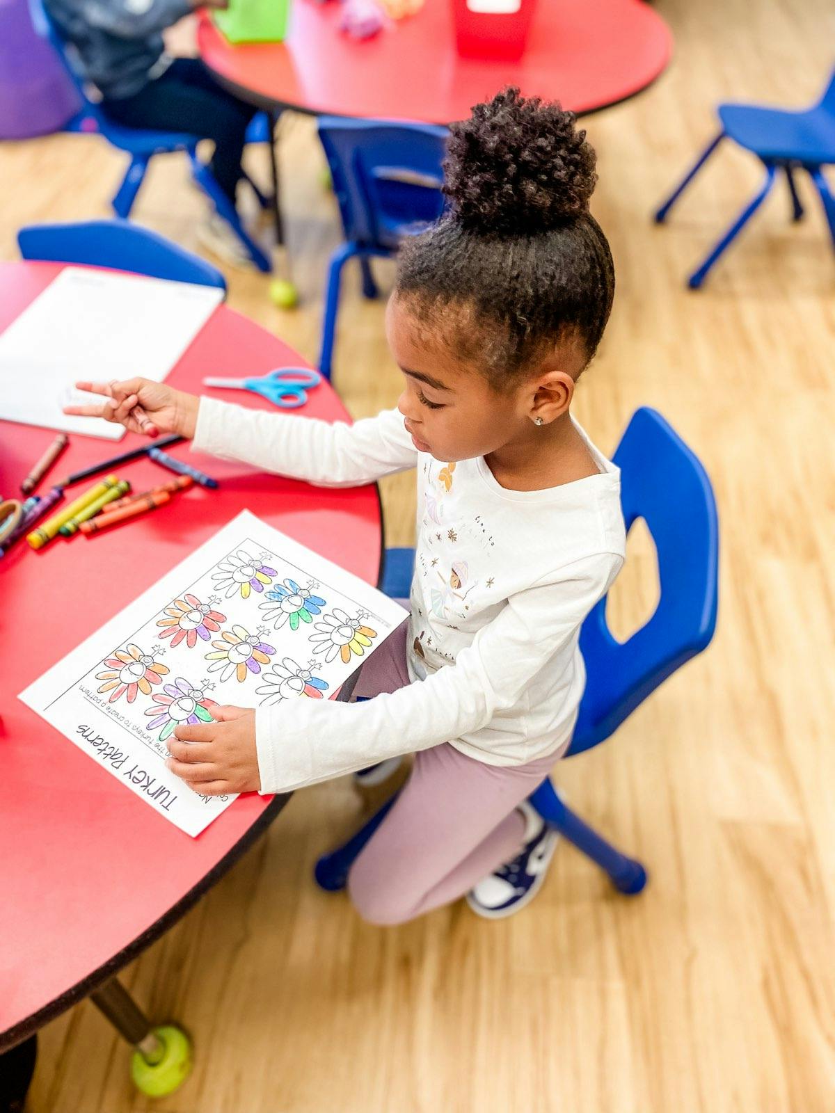 Child coloring a worksheet at a red table in a classroom