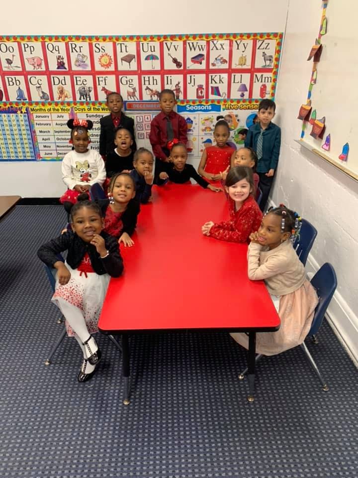 Group of children sitting and standing around a red table in a classroom