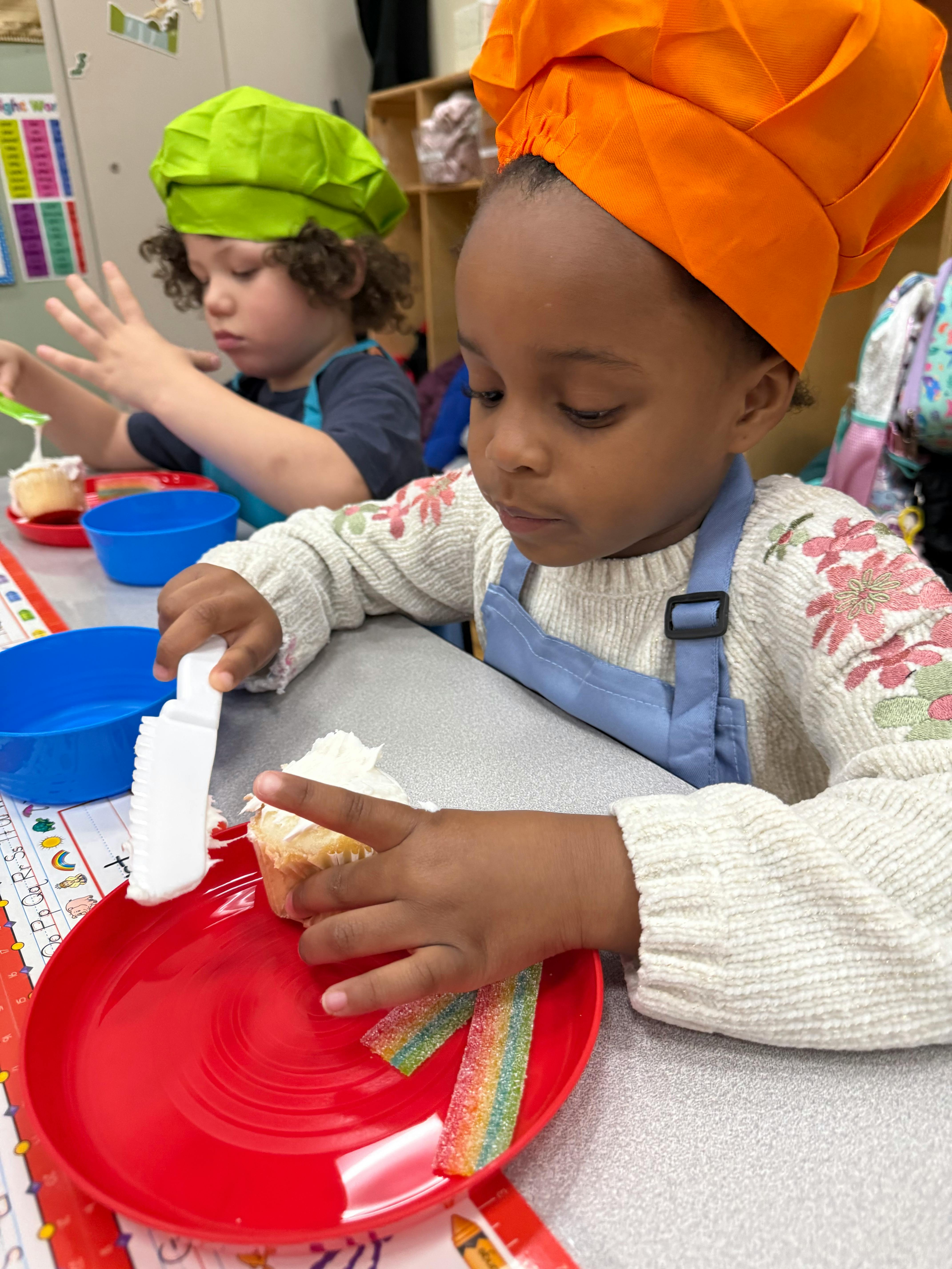 Child in chef hat decorating a cupcake with a knife