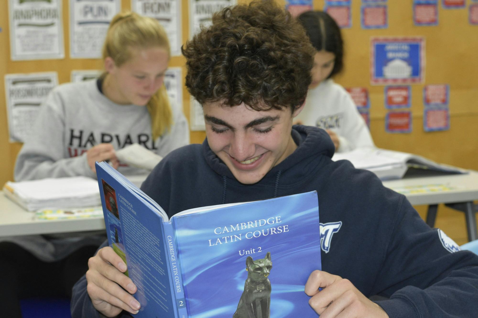 Smiling teen in a classroom reading "Cambridge Latin Course" textbook