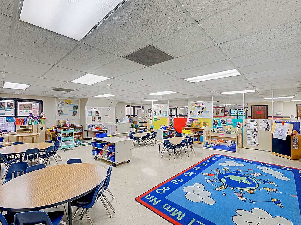 An empty classroom with small tables and chairs, and a colorful rug