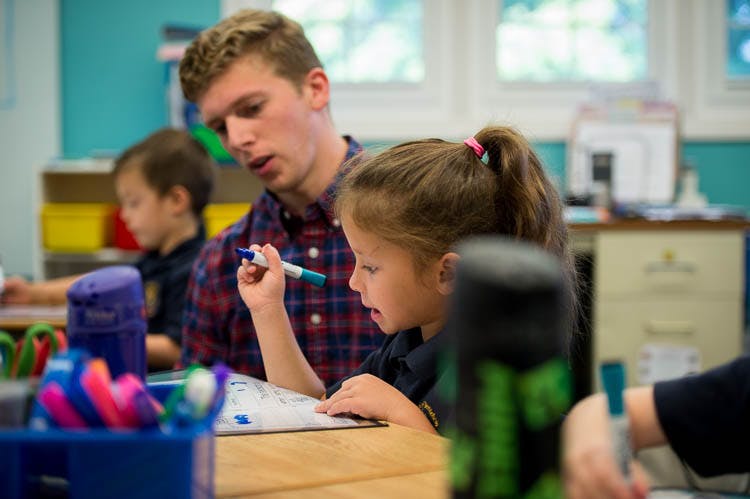 Adult person helping a child write at a desk in a classroom