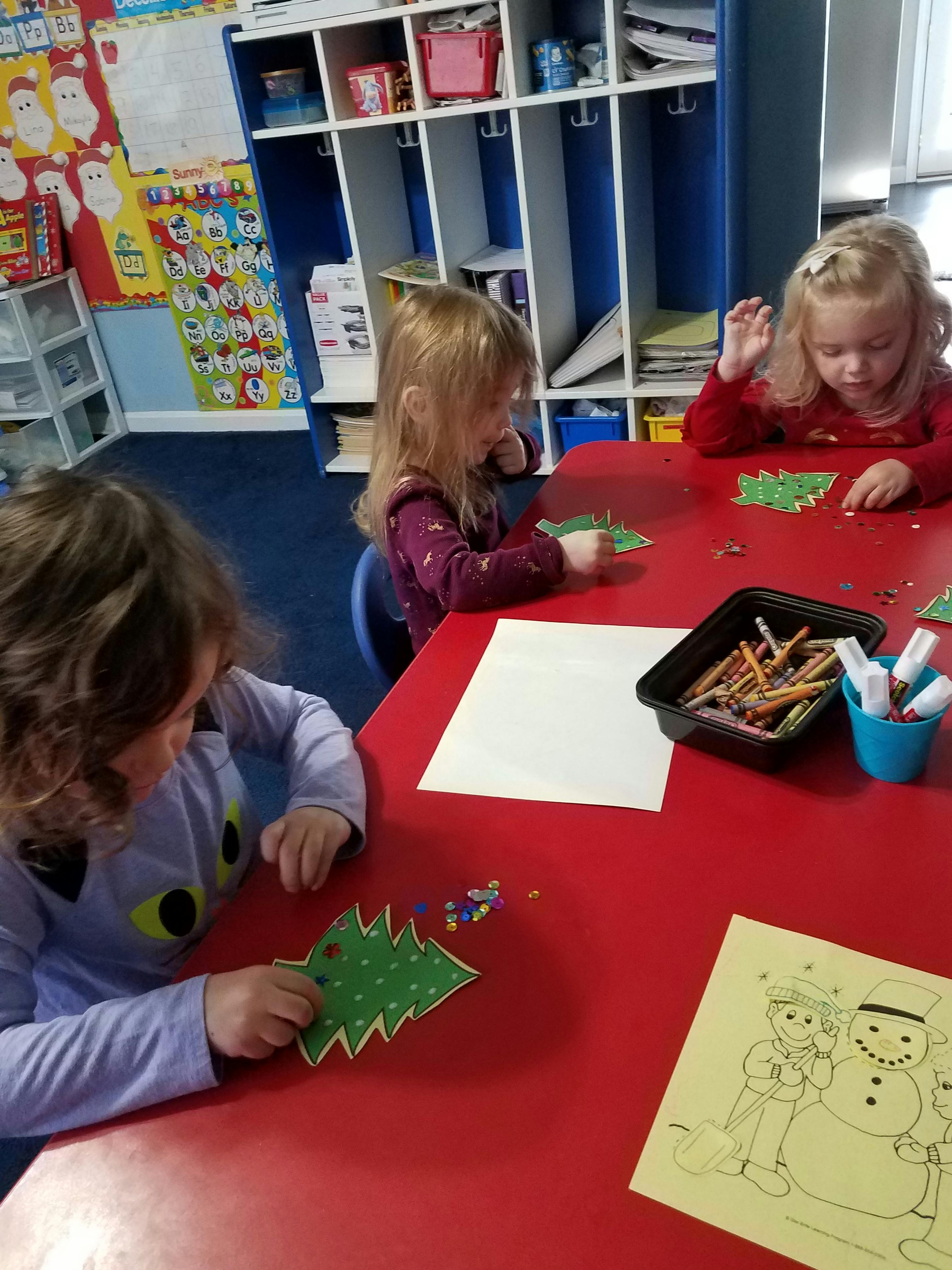 Three children doing crafts at a red table in a classroom