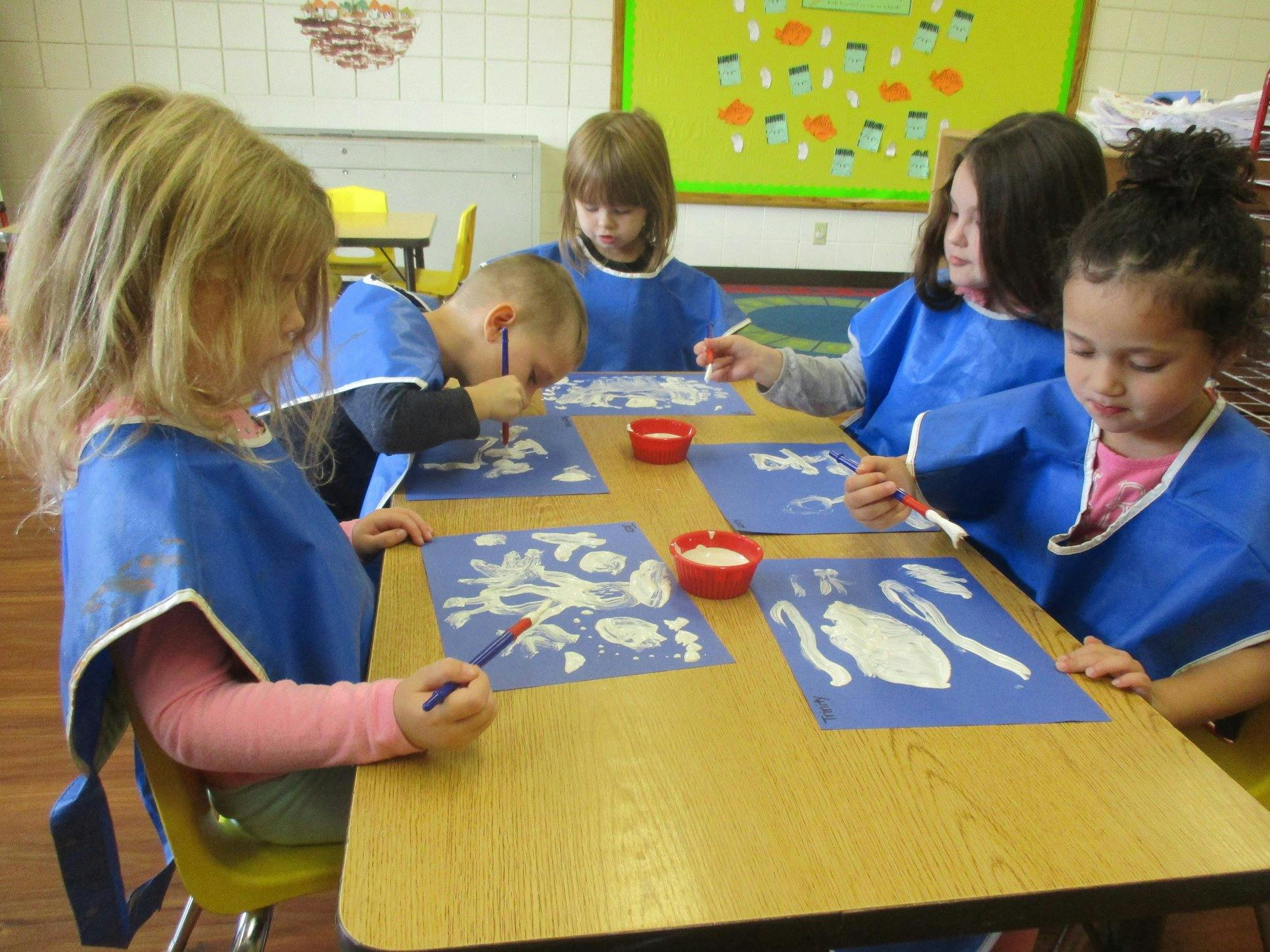 Children in blue smocks painting at a table in a classroom