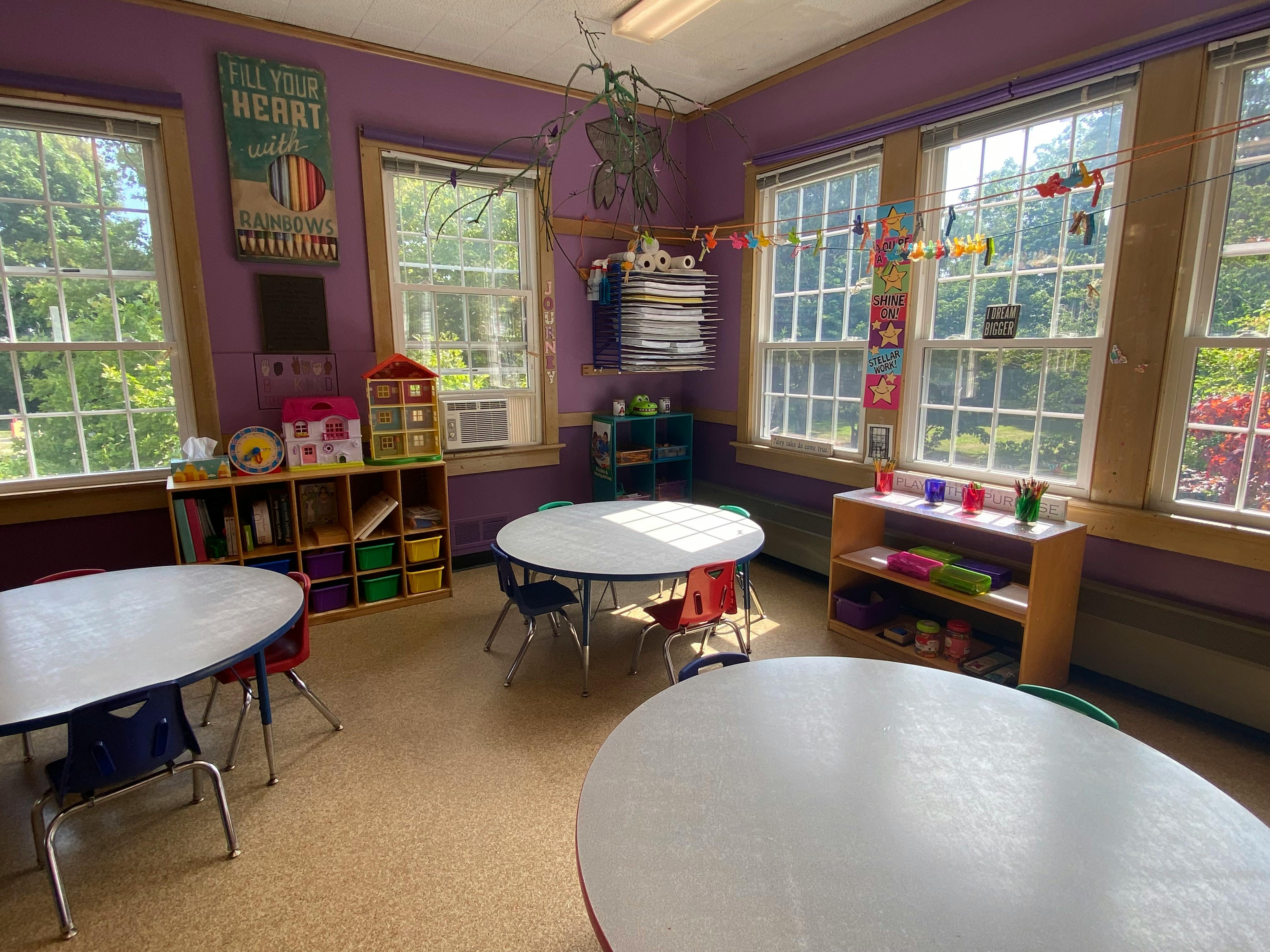 Classroom with small tables and chairs, shelves with supplies, and large windows