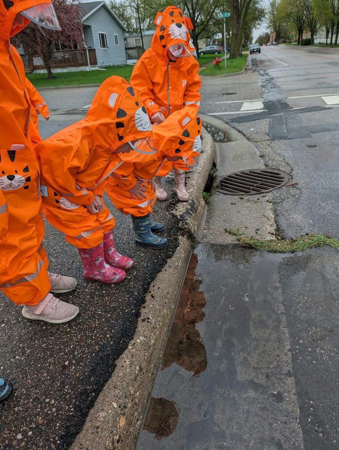 Children in orange rain suits looking into a puddle next to a storm drain